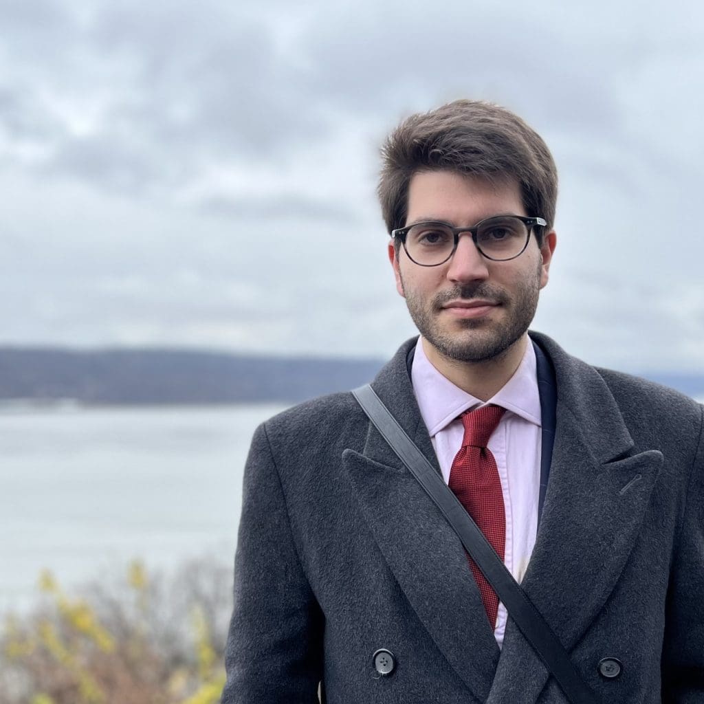 man with brown hair  wearing a suit in glasses standing in front of a body of water