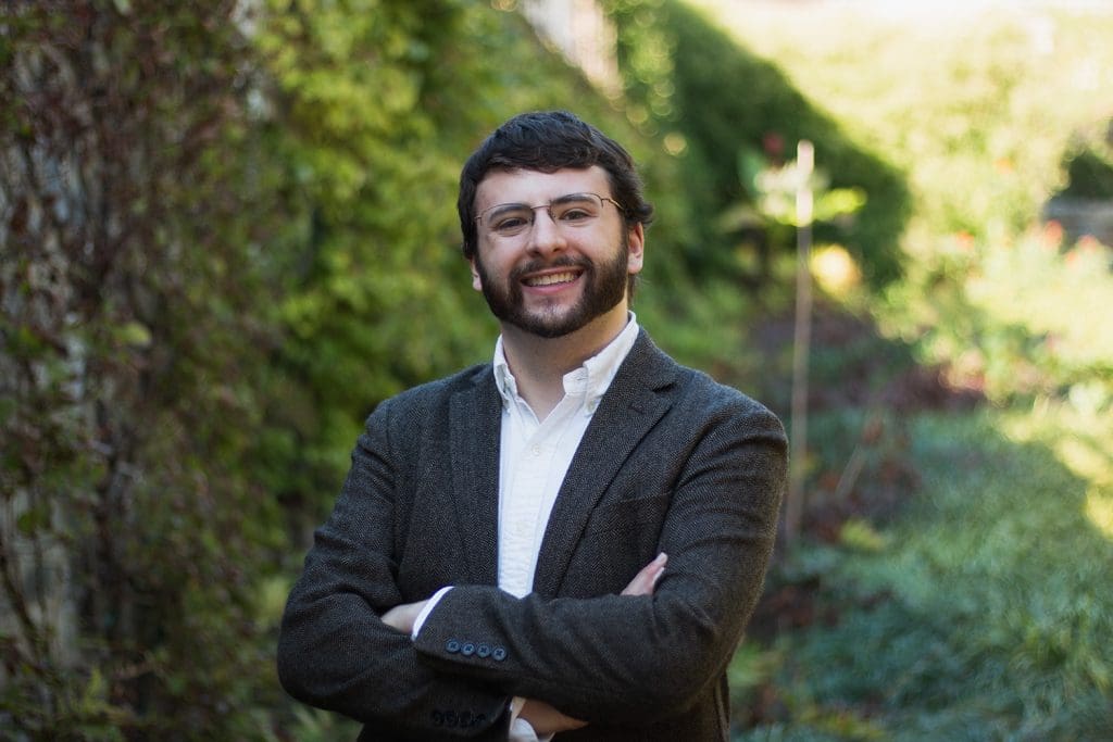 man standing in front of bushes/trees. His arms are crossed, he is wearing glasses and smiling